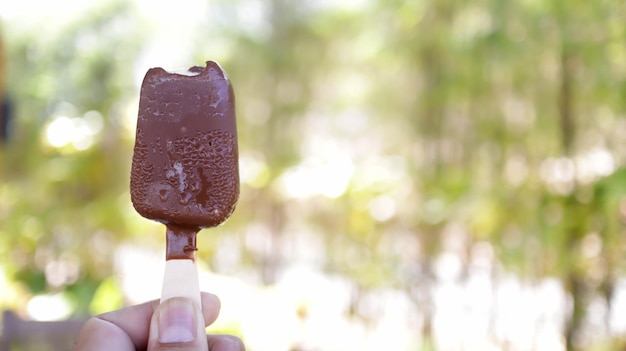 Photo a hand holding bitten common popular dark chocolate ice cream stick or ice bream bar with nature background copy space magnum ice cream