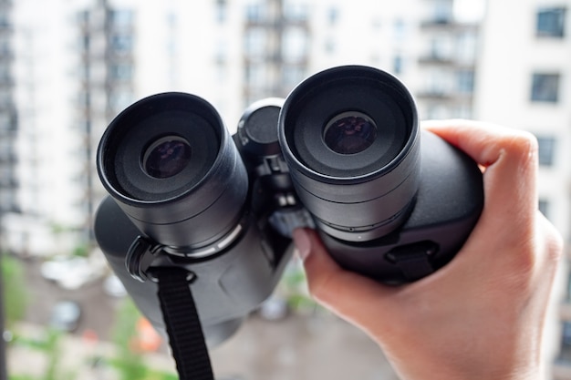 Hand holding black binoculars against a blurred background of a window with a view of the neighboring house for observing neighbors, environment or nature. Selective focus. Closeup view