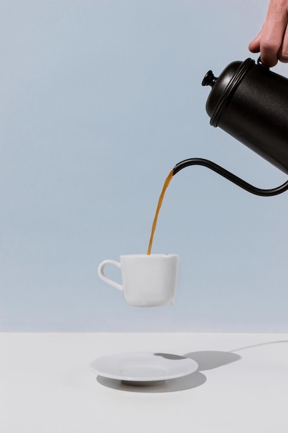 A hand holding black metal pot and pouring coffee into levitating white cup