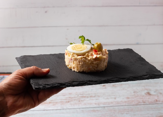 Hand holding a black slate plate with Russian Salad Typical Spanish tapa