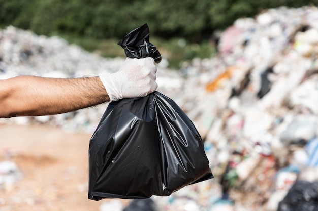 Hand holding a black trash bag in front of a large mountain and a pile of trash. Garbage plastics from industrial and community. World Environment Day.