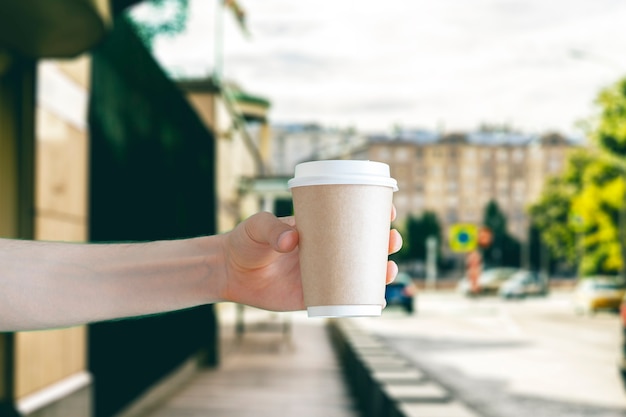 A hand holding a blank coffee paper cup for takeaway outside in the street