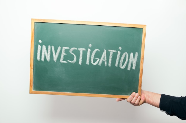 Hand holding a chalkboard with the word INVESTIGATION handwritten in chalk