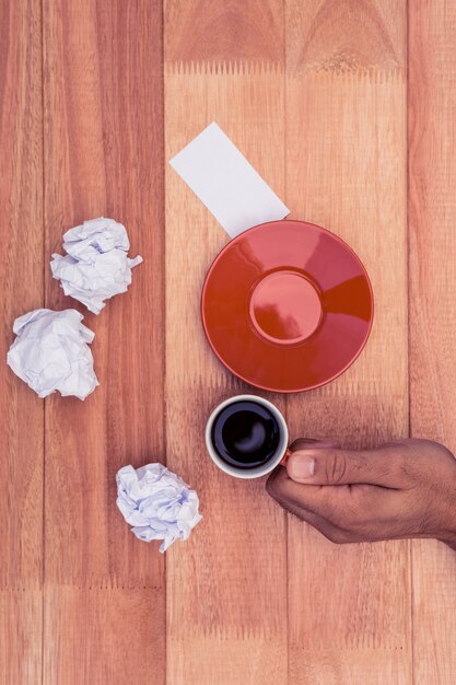 Hand holding coffee cup by crumbled paper and saucer on desk in office 