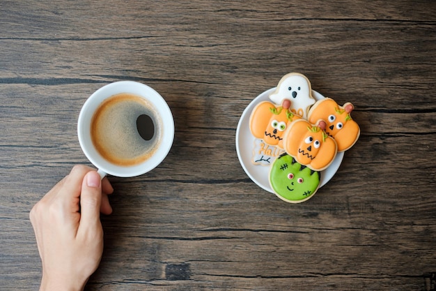 Hand holding coffee cup during eating funny halloween cookies\
happy halloween day trick or threat hello october fall autumn\
traditional party and holiday concept