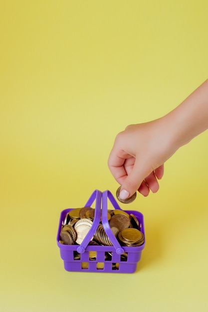 Hand holding a coin with pile of coin in the shopping basket on yellow background