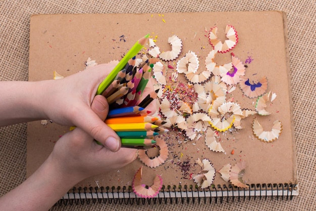Рука держит цветные карандаши над блокнотом с карандашной стружкой