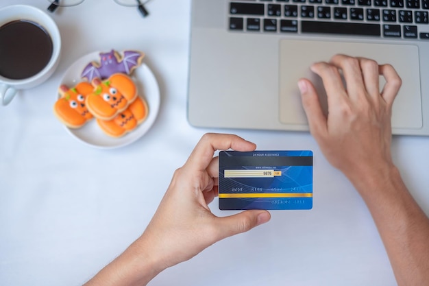 Hand holding credit card for online shopping on laptop during eating Halloween Cookies and coffee Happy Halloween Hello October fall autumn Festive party and holiday concept