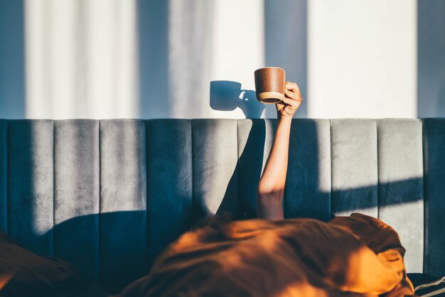 Photo hand holding a cup of coffee while lying on bed. morning concept.