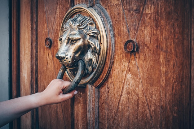 A hand holding a door knocker knocking on a wooden door with a\
metal ring shaped door knocker