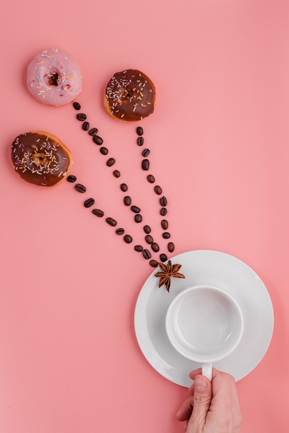 Hand holding empty cup, beans and three doughnut  on pink background