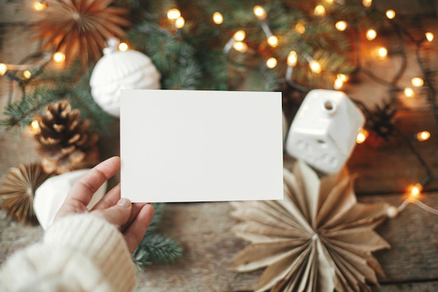 Photo hand holding empty greeting card on background of christmas lights and decor christmas card mockup