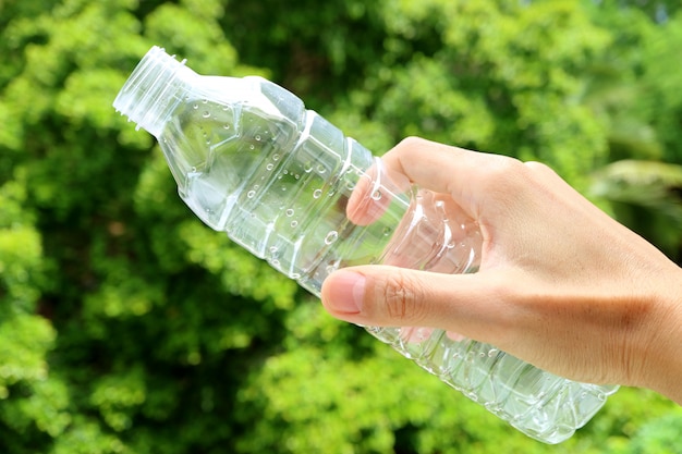 Рука пустую пластиковую бутылку питьевой воды с зеленой листвой на заднем плане