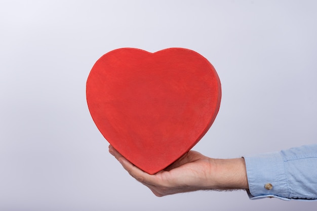 Hand holding festive red box heart shape on white wall. Gift for beloved. Present for Valentine's day