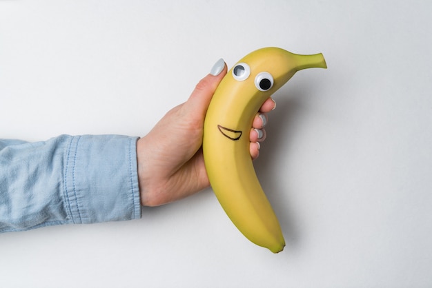 Hand holding fun banana with Googly eyes and smile on white wall