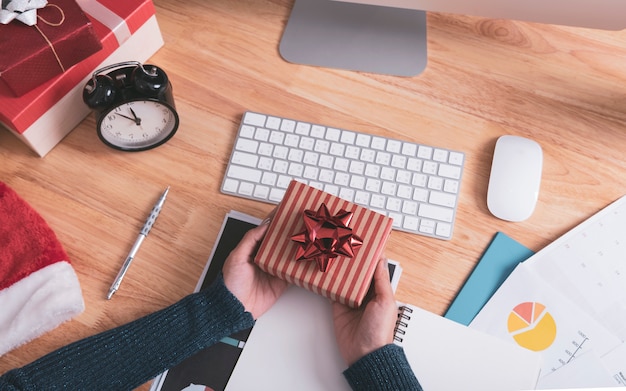 Mano che tiene il contenitore di regalo in festa di natale in ufficio con decorazione di natale sul tavolo.