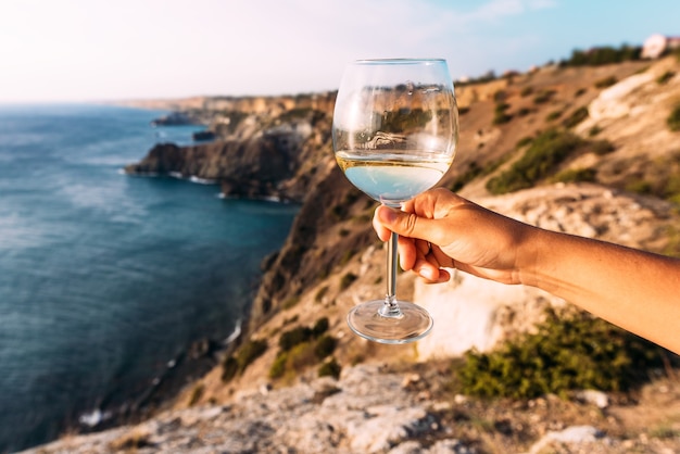 海の上にグラスワインを持っている手。ロマンチックな休暇。海に対してグラスワインを持っている手。海沿いのワインと夕日に出会う。コピースペース