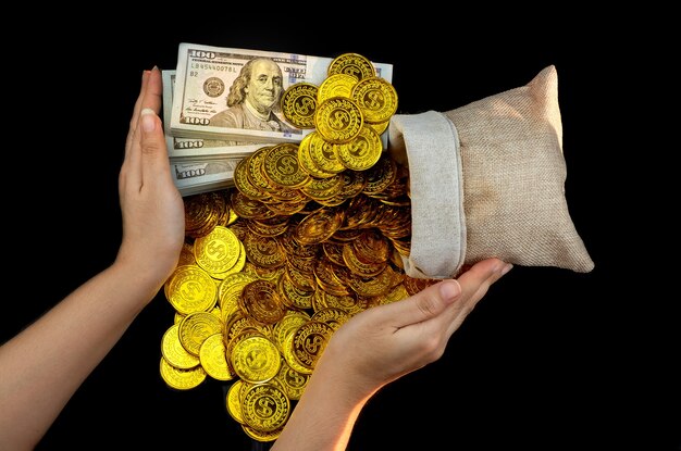 Hand holding gold coin in treasure sack and stack bundles of 100 US dollars banknotes on black background