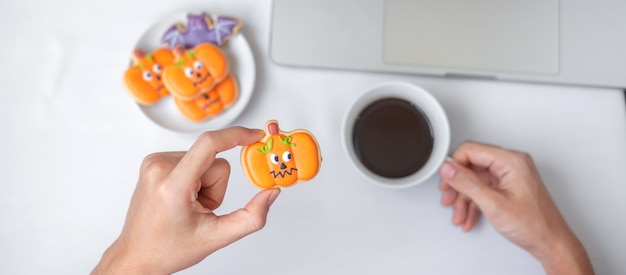 Hand holding Halloween pumpkin Cookie and drinking coffee during using computer laptop. Happy Halloween, online shopping, Hello October, fall autumn, Festive, party and holiday concept