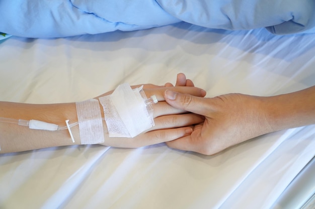 Hand holding the hand of a sick kid and mother on bed were connected to the saline solution in hospi