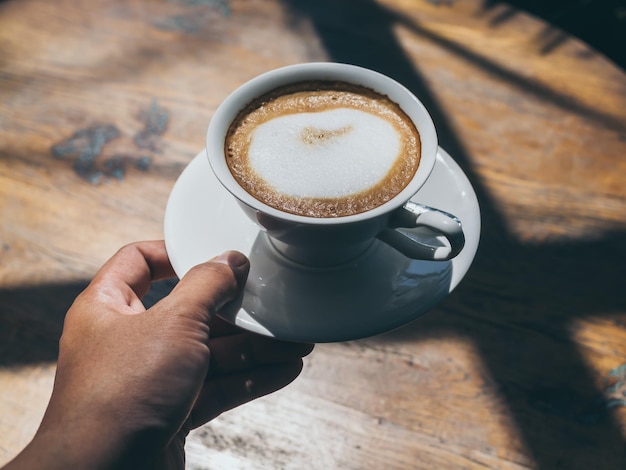 Hand holding hot coffee in round white ceramic cup on wooden table background