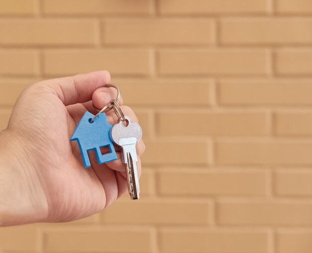 Hand holding a key ring of one key and a mini blue house over a brick background Real State