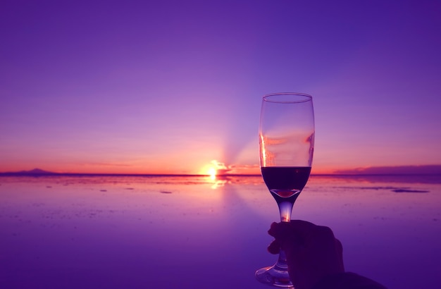 Mano che tiene il bicchiere di liquore contro il tramonto sulle saline di salar de uyuni in bolivia