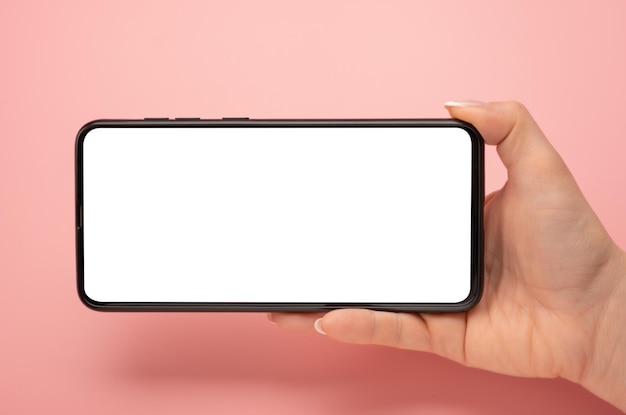 Hand holding mobile phone in horizontal position for mockup watching streaming video on cellphone on pink table background Close up of horizontal black smartphone with blank screen in woman hands