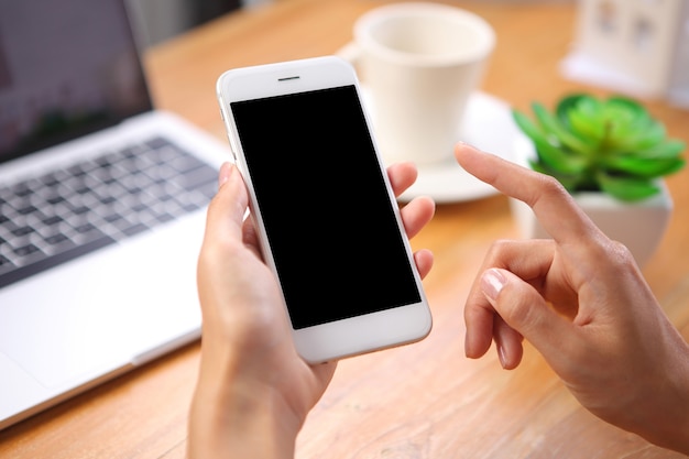 Hand holding mockup smartphone with laptop and office stationery on wooden desk