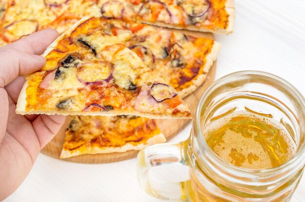 Hand holding piece of pizza over table with pizza and glass of beer