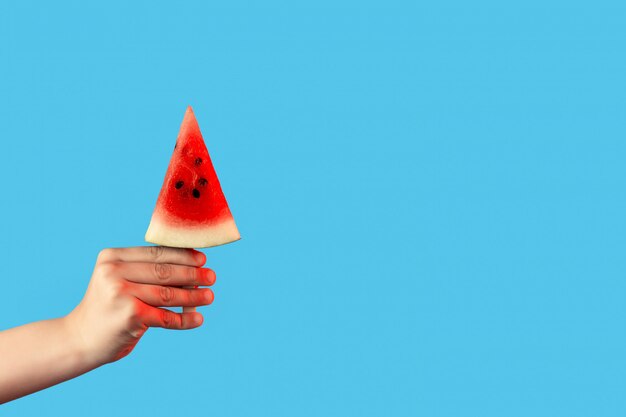 Hand holding a piece of watermelon on a stick like ice cream on blue 