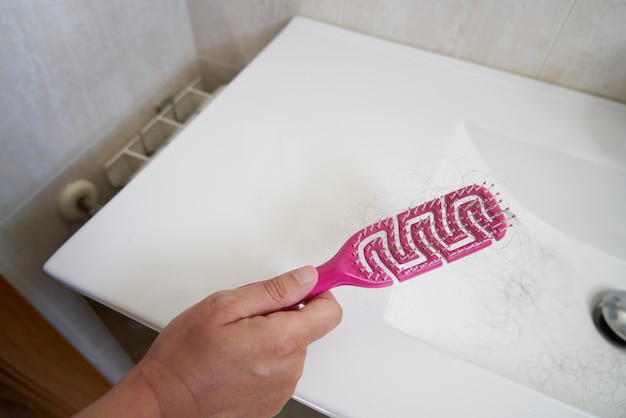 Foto mano che tiene un pettine rosa con peli aggrovigliati a causa di un problema di perdita di capelli in un lavandino