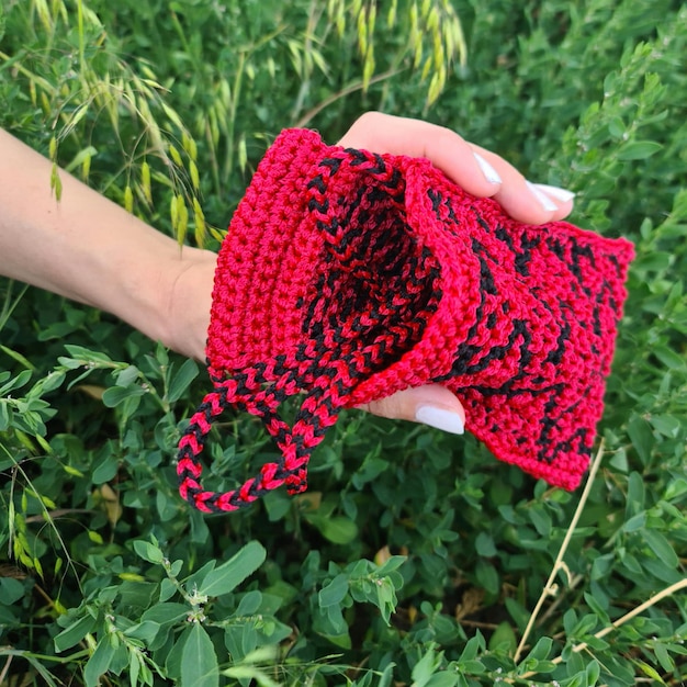 A hand holding a red and black knitted cloth in a field of green plants