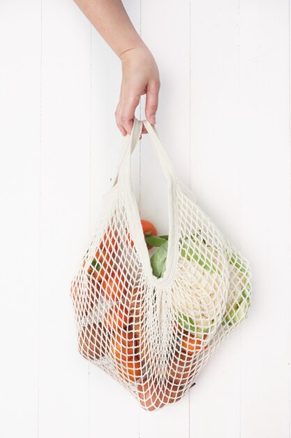 Hand holding a reusable mesh bag filled with fruits and vegetables on white wooden background