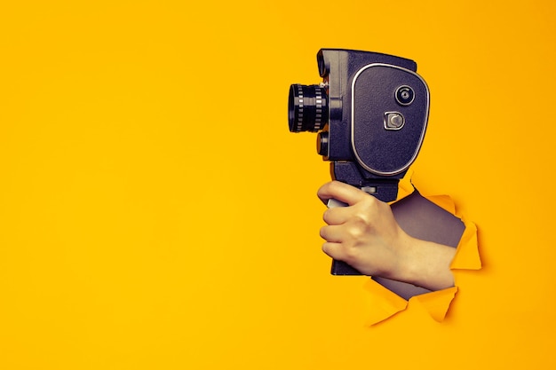 Hand holding small video old retro black camera 8 mm in paper torn hole in yellow background with copy space