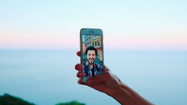 Foto mano che tiene smartphone durante la videochiamata con l'uomo di fronte al mare bellissimo paesaggio nel ba...