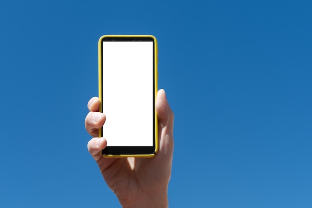 Hand holding a smartphone vertically outdoors with sky and greenery in the background a sunny day