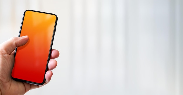 Hand holding a smartphone with blank red and orange screen Office background Horizontal banner