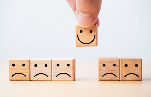 Hand holding Smile face print screen on wooden cube block among sadness face for customer service evaluation and emotion mindset concept.