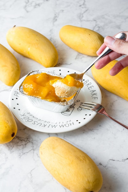 La mano che tiene un cucchiaio ha raccolto la torta al formaggio al mango la torta al formaggio al mango è confezionata in un foglio di alluminio quadrato in un piatto bianco su una torta da tavolo di marmo in un bar o in una panetteria