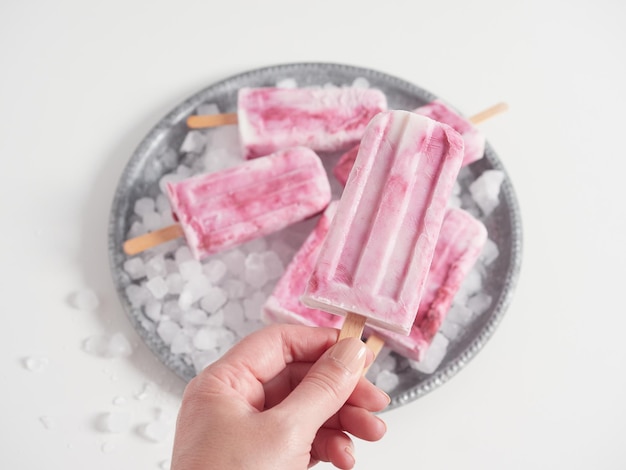 Foto mano che tiene il bastoncino di un ghiacciolo fatto in casa con yogurt e gelato alla fragola sullo sfondo altri su ghiaccio in un vassoio d'argento rotondo