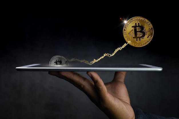 Hand holding tablet with bitcoins and graph coming out of it on dark background