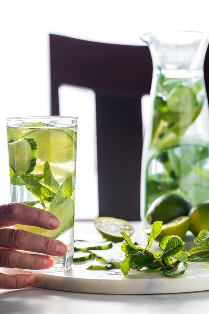 A hand holding a tall glass of infused cucumber water and a full pitcher beside