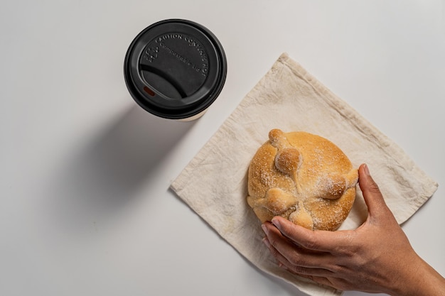 Hand holding a traditional homemade Mexican bread of the dead next to a cup of coffee on white table