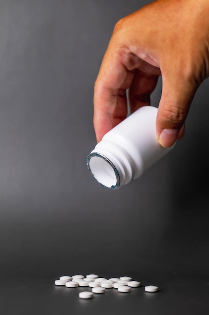 Hand holding unlabeled white plastic pill bottle open cap Poured out several small white pills come out below in black background