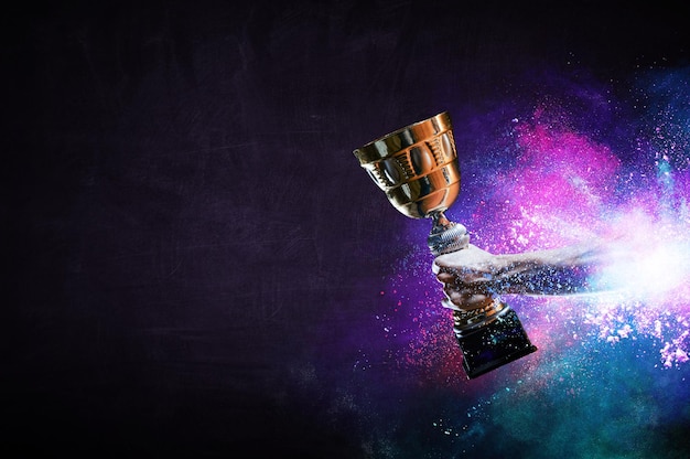 Hand holding up a gold trophy cup against dark background. Mixed media