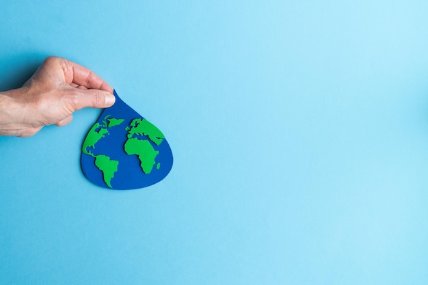 Photo hand holding a water drop cut out of paper with world map inside on blue background international earth day copy space