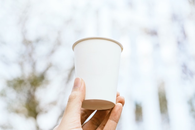 Hand holding white blank takeaway paper, carton or cardboard coffee cup with blur.