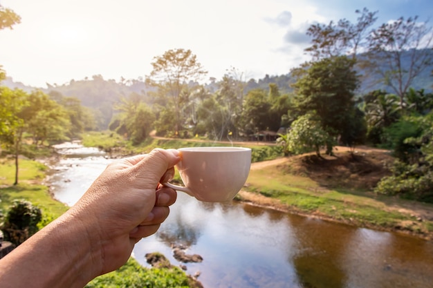 Hand holding a white cup of hot espresso coffee mugs and nature view of the mountain landscape in the morning with sunlight