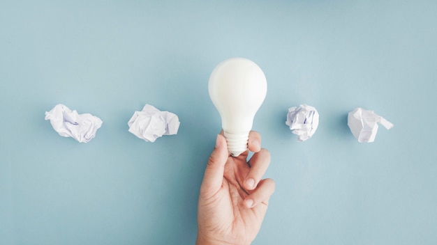 Photo hand holding white light bulb with crumpled paper balls on gray background